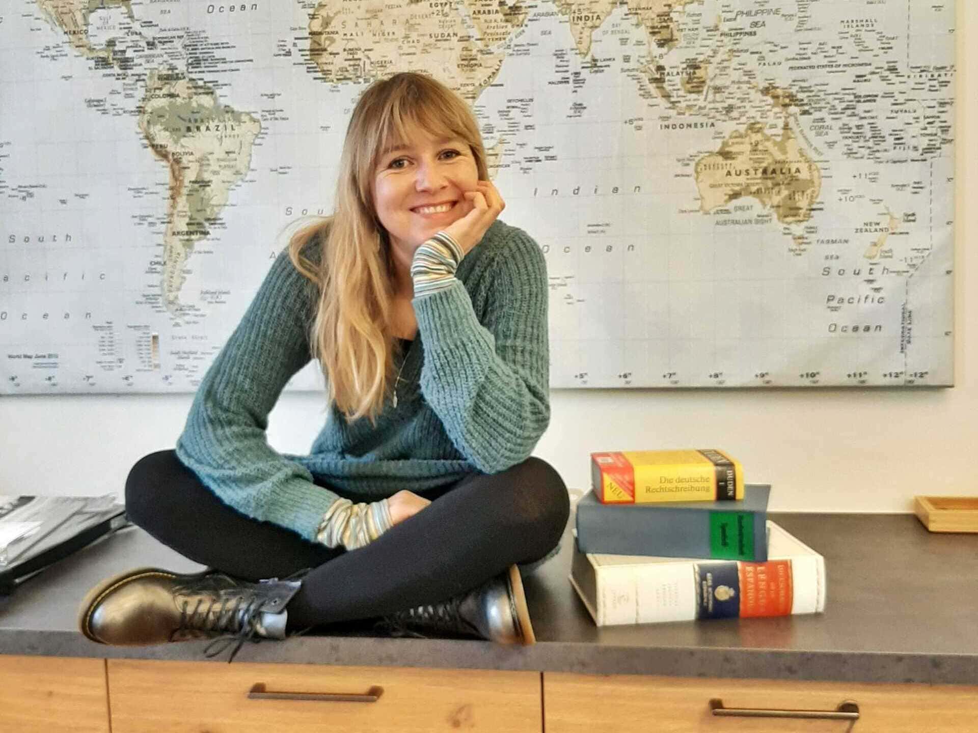 Mitglied vom FrauenRaum Netzwerk Fabienne Feuz von Sprachart sitzt auf einer Kommode, neben ihr einige Büche r und hinter ihr an der Wand ist die Weltkarte zusehen.